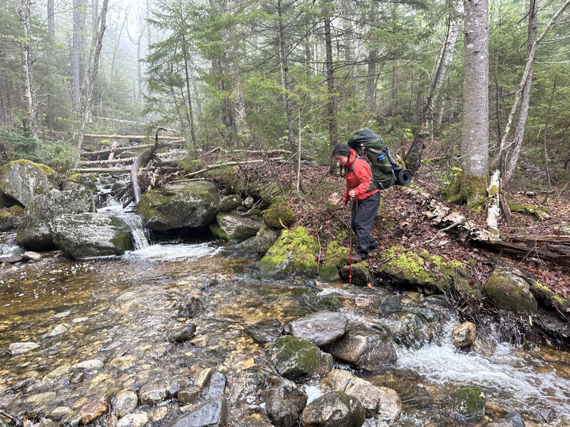 A person with a large backpack is carefully crossing a rocky stream in a forest. The water is clear and flowing rapidly over the rocks. Moss covers many of the rocks and fallen leaves are scattered around. There's a small wooden bridge in the background, and the trees are dense, creating a misty atmosphere.